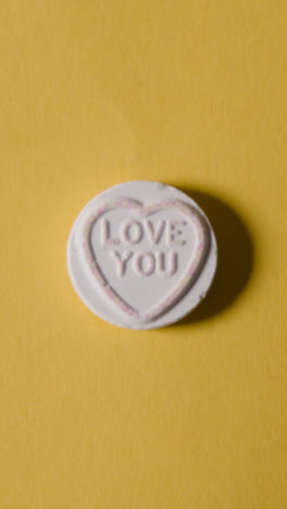 Vertical-Video-Close-Up-Of-Vertical-Video-Of-Hand-Holding-Heart-Candy-With-In-Love-Message-On-Yellow-Background